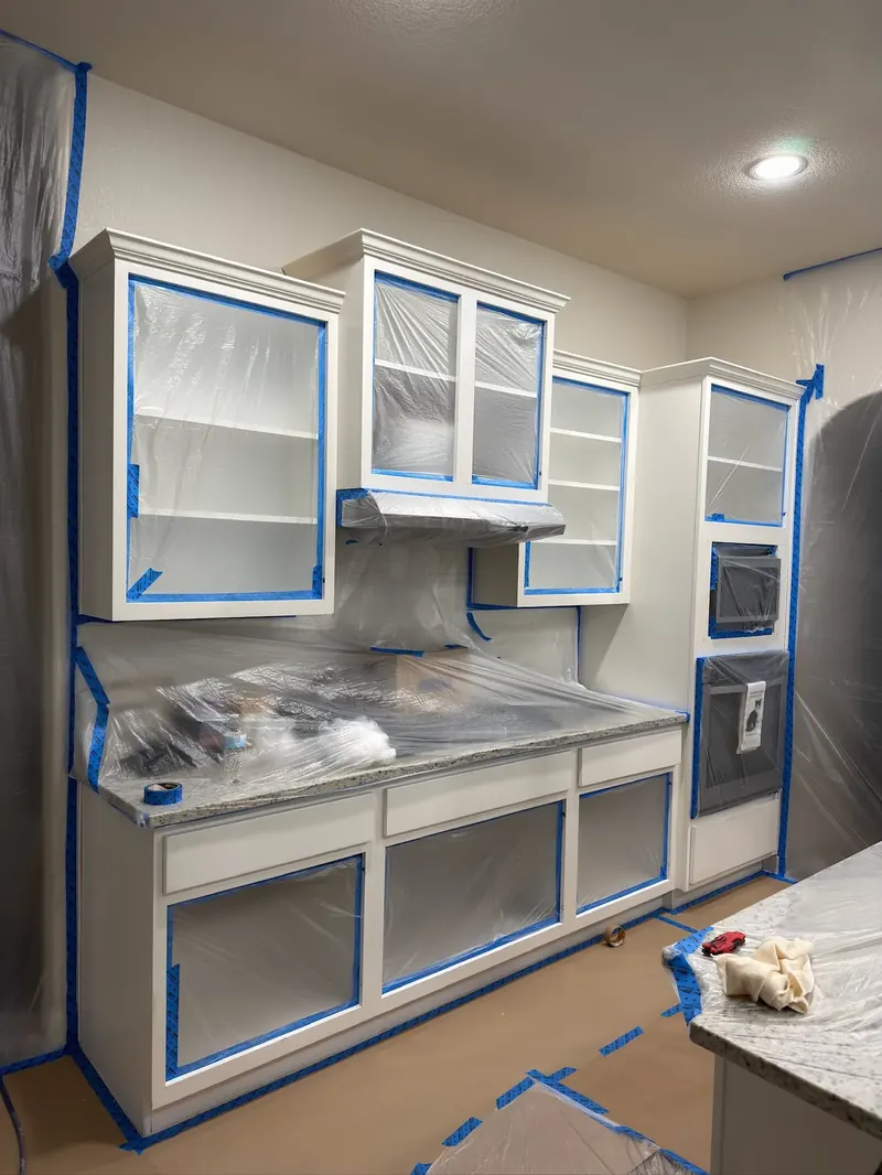 Kitchen cabinets fully masked with plastic and blue tape, doors off, before spray painting, V&S Services Austin