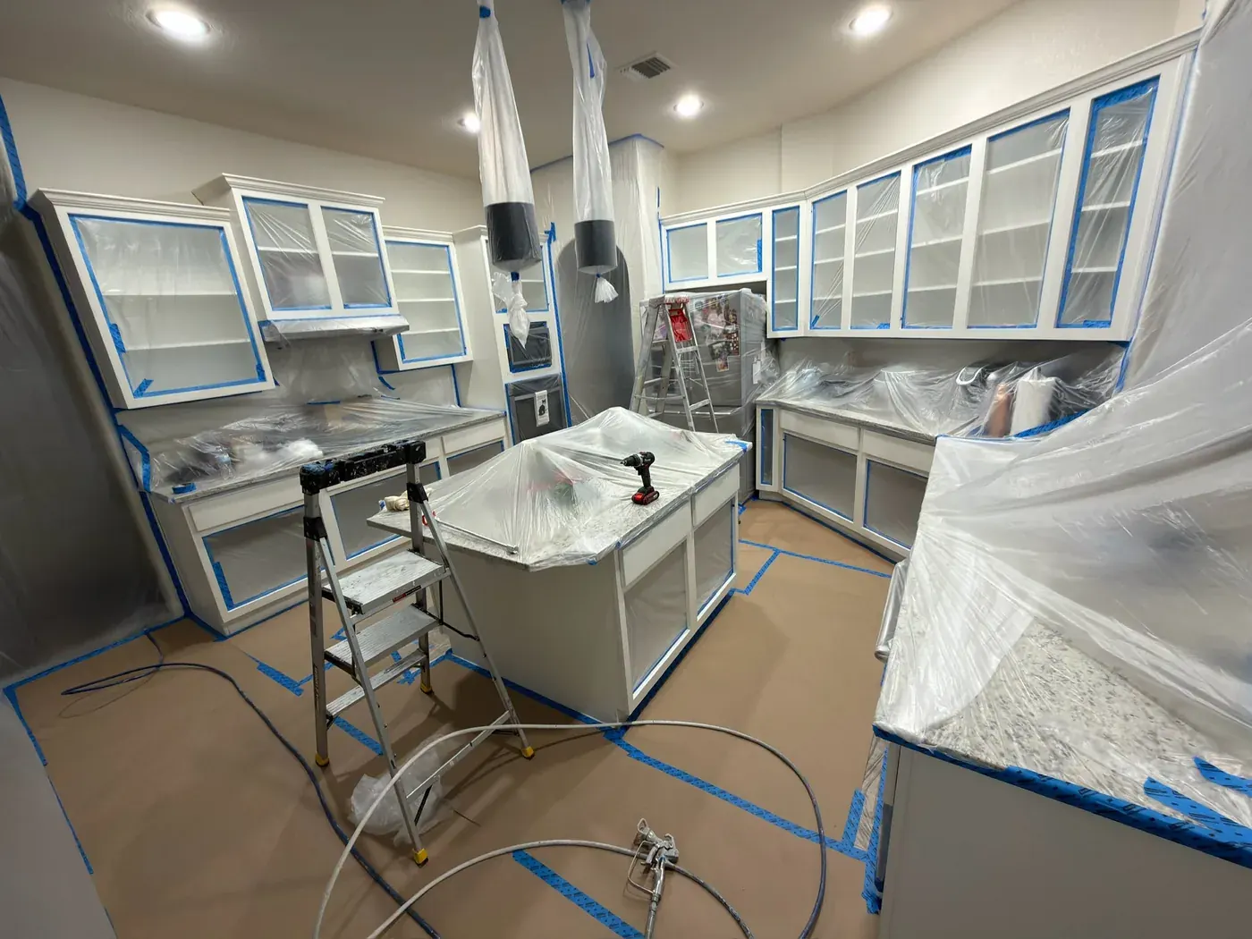Kitchen during cabinet spray painting — full containment with plastic sheeting and airless sprayer, V&S Services Austin